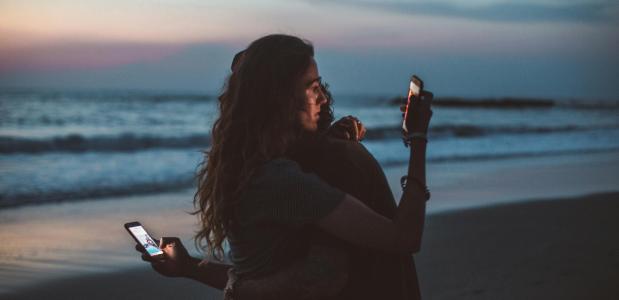 Een man en een vrouw op een strand omhelzen elkaar terwijl ze ondertussen afgeleid op hun telefoons kijken.