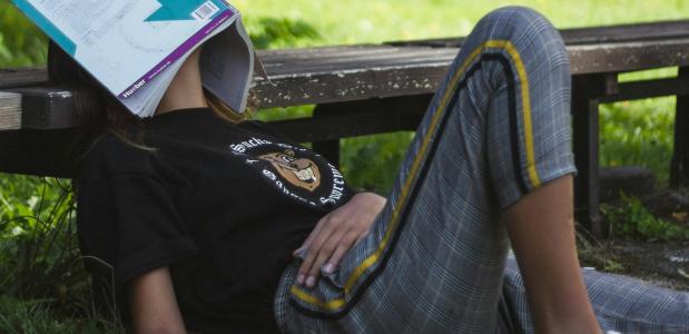 Een vrouw ligt met haar achterhoofd op een bankje en een geopend boek over haar gezicht heen