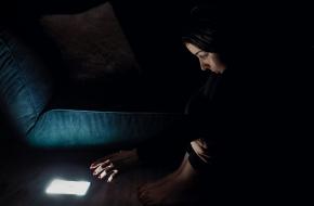 Jonge vrouw reikt met haar hand naar een oplichtende telefoon in een donkere ruimte